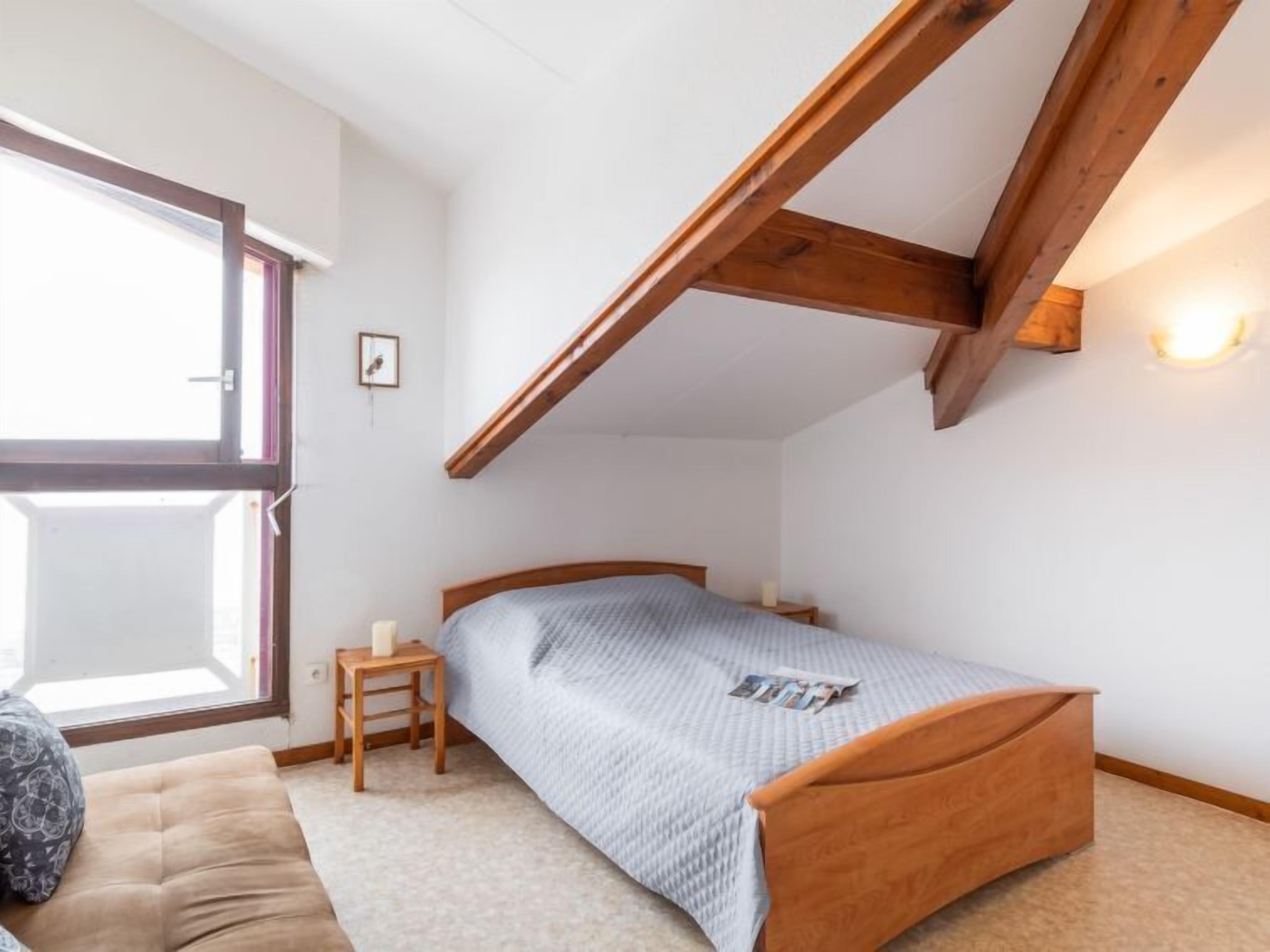 Ein Schlafzimmer mit schrägen Wänden, einem Bett mit blauer Bettdecke, einem kleinen Nachttisch mit Kerze und einem Fenster mit Blick.