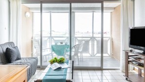 A modern living room with a sofa, a coffee table with a book and flowers, a TV stand, and a balcony with a table and chairs. - Belvédère by Interhome (Mimizan)