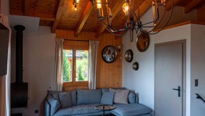 Ett mysigt vardagsrum med ett träpanelat tak, en inbyggd vedkamin, en grå soffa och ett stort fönster med gardiner.