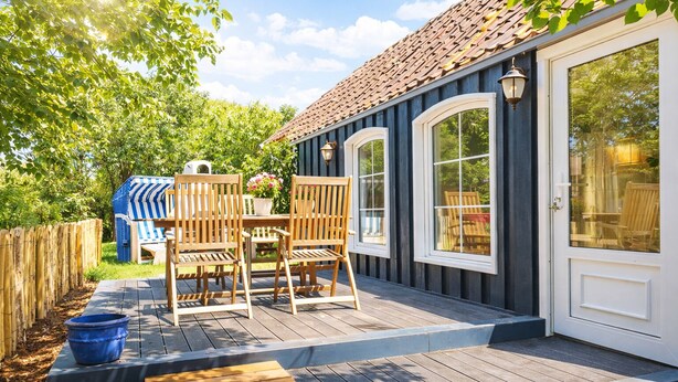 Een houten terras met buitenmeubilair, een overdekte patio en een huis met een donkere gevel en een pannendak.