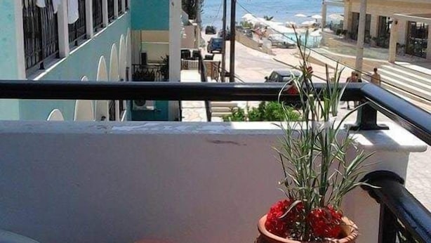 A balcony with a table set for two, a potted plant, and a view of the sea and buildings. - Hotel Asimina (Zakynthos)
