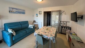 A compact living space with a sofa, a dining table, a TV mounted on the wall, and a small kitchenette area.