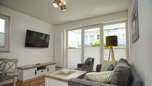 Ein modernes Wohnzimmer mit grauem Sofa, einem Holztisch, einem an der Wand montierten Fernseher und einem Fenster mit Jalousien.