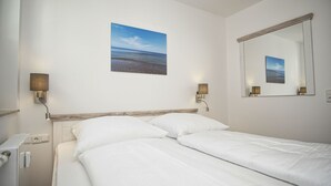 Ein Hotelzimmer mit Bett, Nachttisch, Lampe, einem an der Wand hängenden Bild von der See und einem Spiegel.