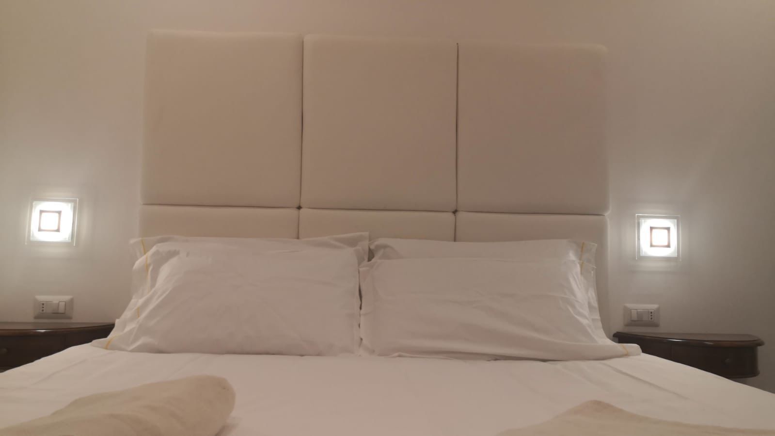 A neatly made bed with white linens and two pillows, a padded headboard, and bedside lamps on either side.