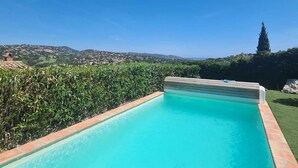 A rectangular swimming pool with a clear blue water, surrounded by a brick border, set against a backdrop of a lush green hedge and a distant view of a hilly landscape under a clear blue sky. - Villa Avec Apercu MER ET Piscine Privee (Sainte-Maxime)