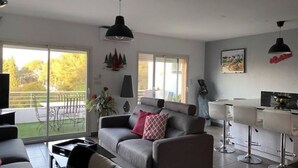 Un salon moderne avec un canapé gris, une table basse et un téléviseur. Il y a un balcon avec vue sur les arbres et un espace patio aménagé avec du mobilier d’extérieur.