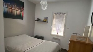 Une chambre à coucher comprenant un lit, une table de chevet, une commode, une fenêtre avec des stores et un tableau représentant un paysage urbain accroché au mur.