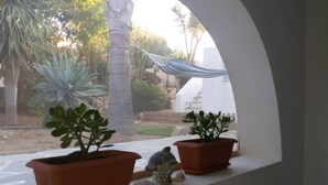 A window with two terracotta pots containing plants, a small figurine, and a view of a garden with palm trees and a blue canopy. - Riviera paradise-unique villa with private pool (Καμάρι)