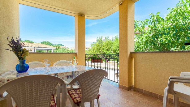 Un patio esterno coperto con tavolo e sedie, un vaso con fiori e vista su aree verdi ed edifici.