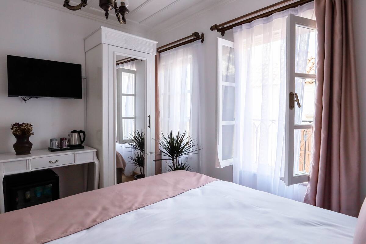 A hotel room with a bed, a desk with a flat-screen TV, a small refrigerator, and a window with curtains.