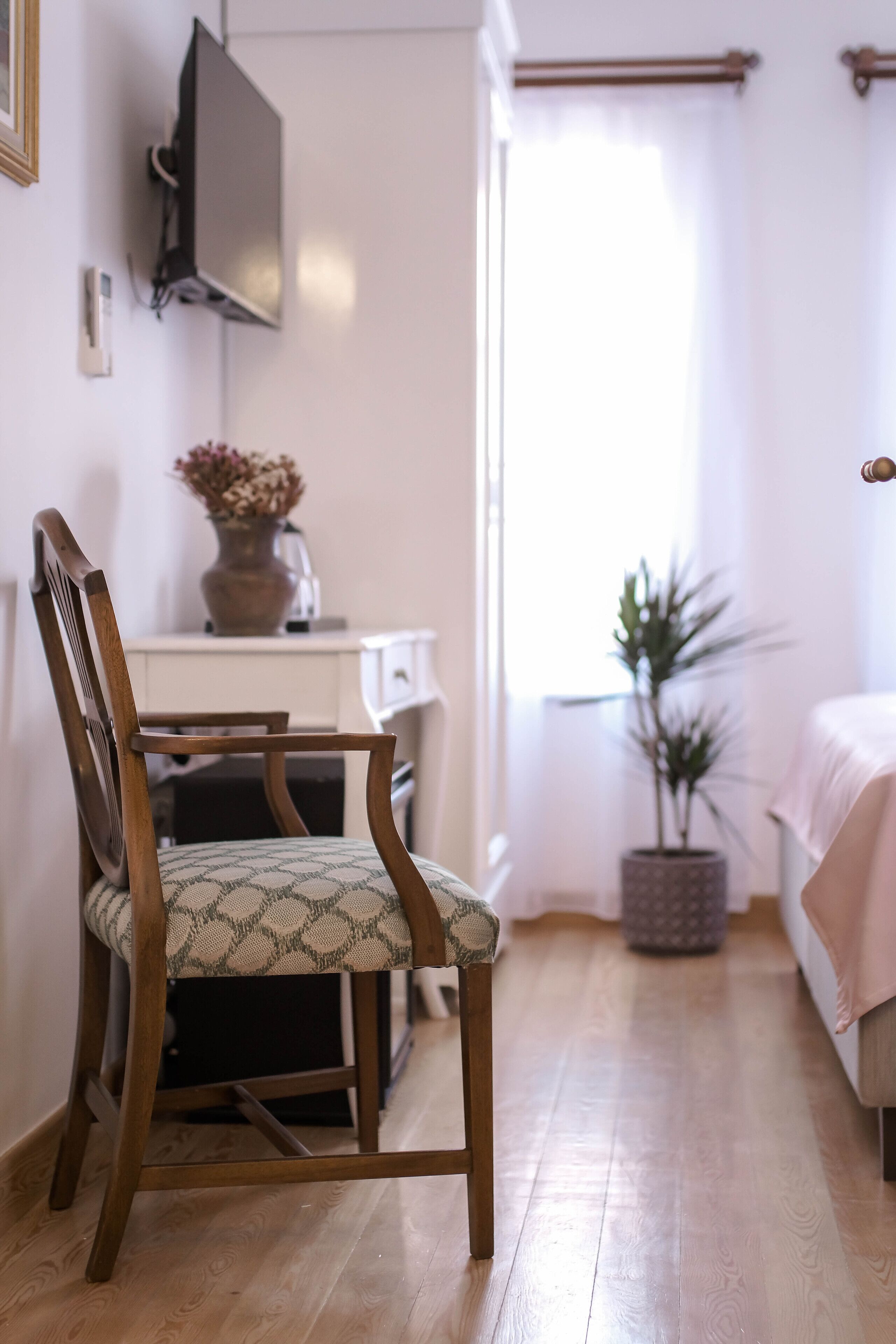 A wooden chair with a patterned cushion in a room with a wooden floor, a white cabinet, a television mounted on the wall, a potted plant, and a bed with a pink blanket.