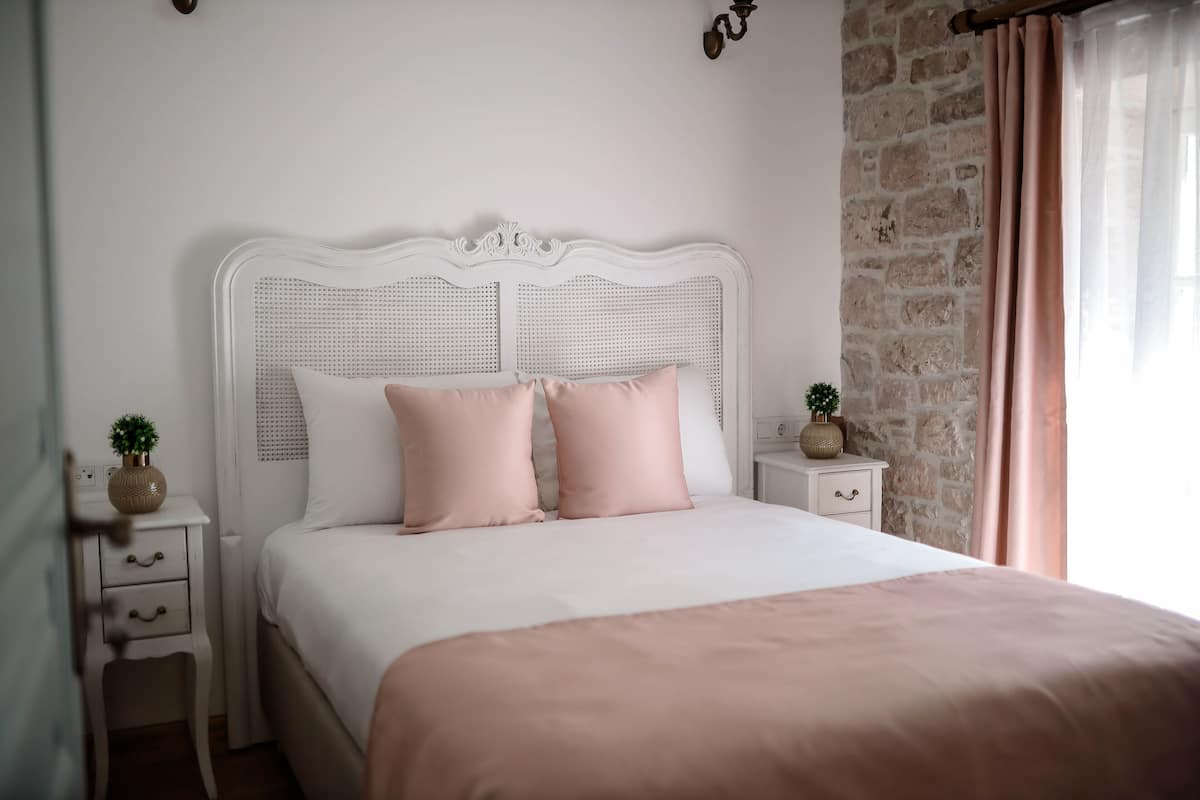 A neatly made bed with a white headboard and two pink pillows, a small bedside table with a plant, and a stone wall to the right.