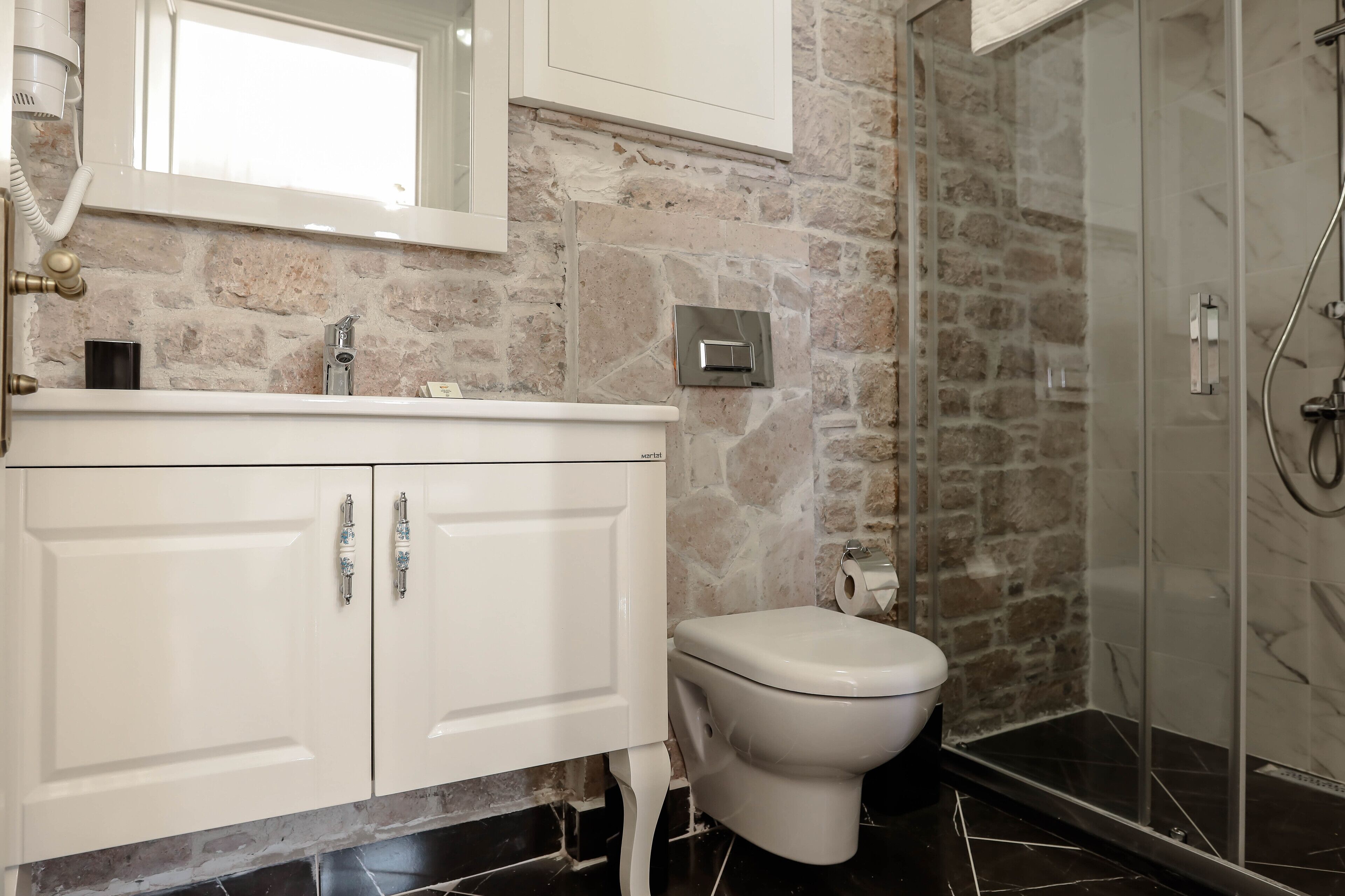A bathroom with a stone wall, a white sink with a mirror, a toilet, and a glass-enclosed shower area.