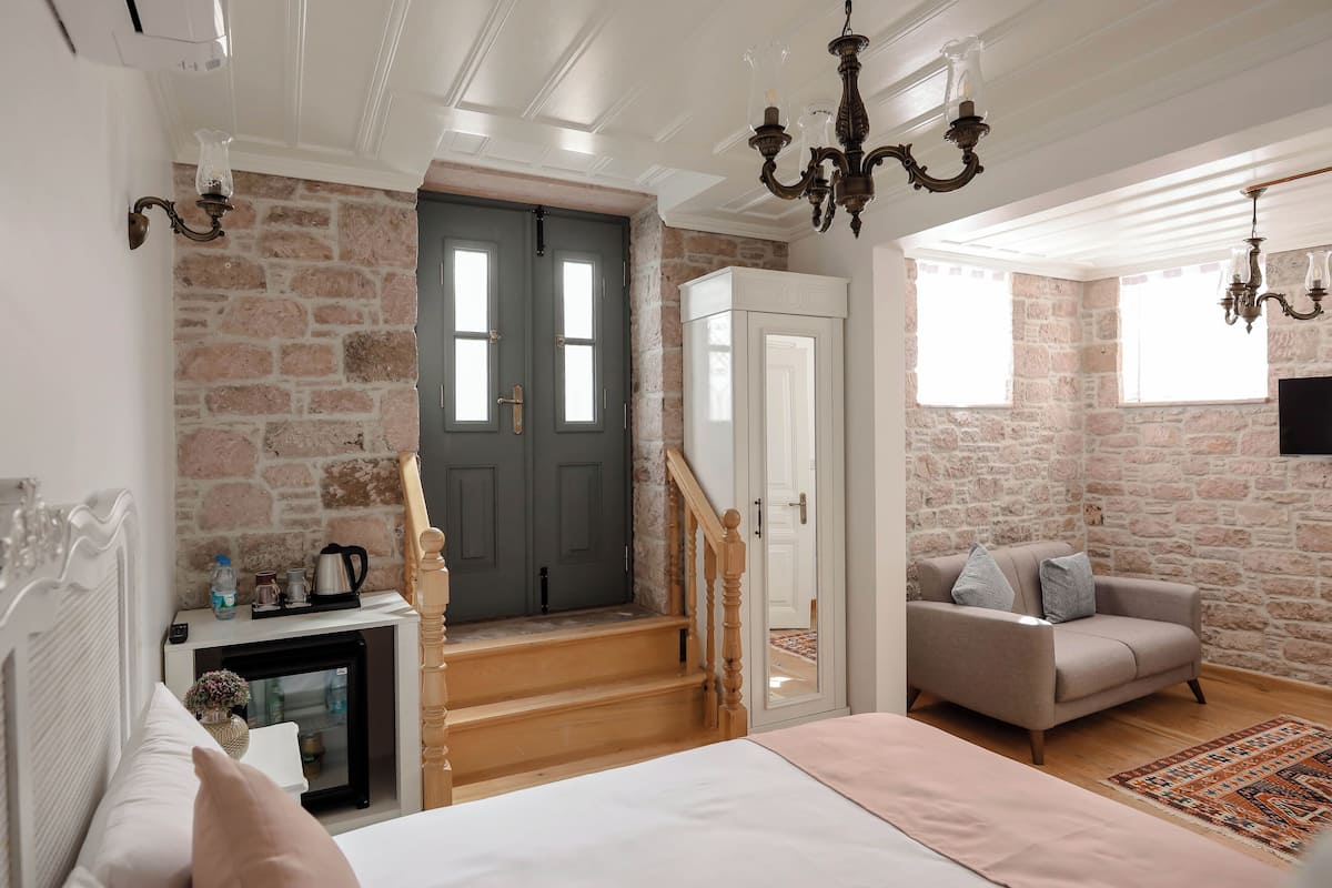 A bedroom with a bed, a dresser, a nightstand, a fireplace, a sofa, a staircase, and a stone wall.