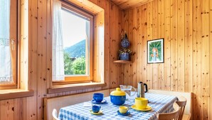 L’intérieur d’un chalet en bois comprend une table à manger dressée pour quatre personnes, une fenêtre donnant sur un paysage verdoyant et une étagère sur laquelle est posée une photo encadrée.
