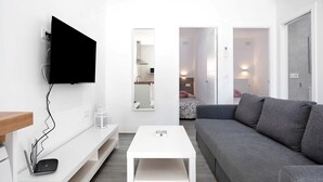 Un salon moderne avec un téléviseur à écran plat fixé au mur, un canapé gris, une table basse blanche et une console blanche sur laquelle sont posés un ordinateur portable et une petite enceinte.