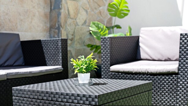 Un espace de détente extérieur aménagé avec des meubles en osier, comprenant une table carrée sur laquelle est posée une plante en pot.