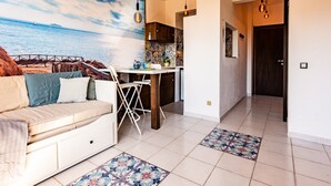 A living room with a sofa, a dining table, and chairs. There is a kitchen area with a tiled backsplash and a door leading to another room. - Studio 'Mono Le Lecce 38B' with Pool and Balcony (Capoliveri)