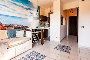 A living room with a sofa, a dining table, and chairs. There is a kitchen area with a tiled backsplash and a door leading to another room.