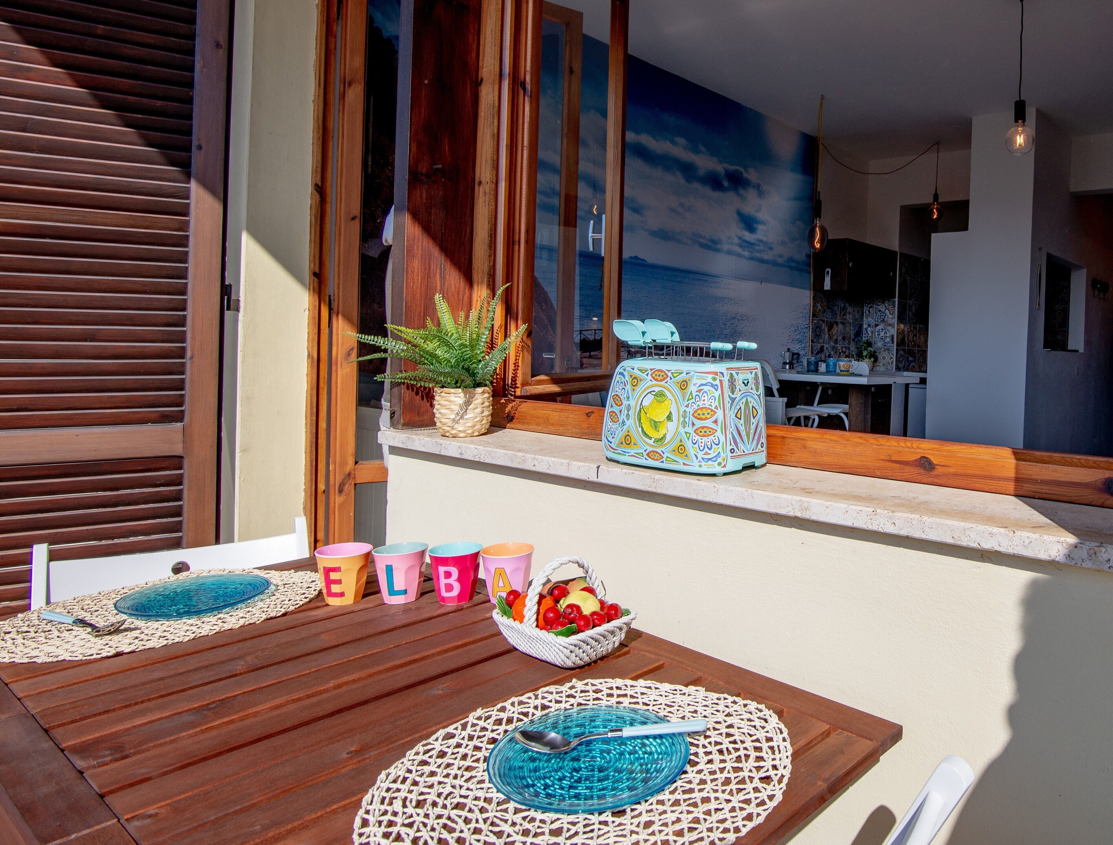 Une terrasse en bois avec une table, des tasses colorées, un panier de fruits et une fresque décorative sur le mur.