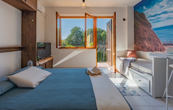 A bedroom with a bed, a desk, a TV, and a balcony with a view of the sea.