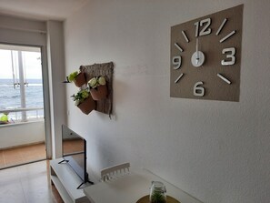 A room with a large wall clock, a window with a view of the sea, and a table with a plant.
