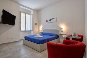 Une chambre à coucher avec un lit, une télévision, une fenêtre, un tableau et un fauteuil rouge.