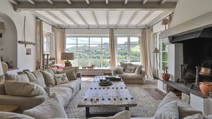 Ein geräumiges Wohnzimmer mit einem großen Holztisch, beigefarbenen Sofas, einem Kamin und großen Fenstern mit Blick auf die Landschaft.