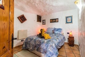 A bedroom with a bed, a wooden door, a wall-mounted heater, and a small table with a lamp. - Vacation Home "Silence Cave Rural House" with Moutain View, Terrace & Wi-Fi (Artenara)
