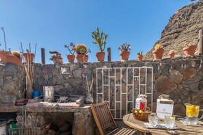 A stone wall with potted plants, a grill, and a table with drinks and a basket. - Vacation Home "Silence Cave Rural House" with Moutain View, Terrace & Wi-Fi (Artenara)