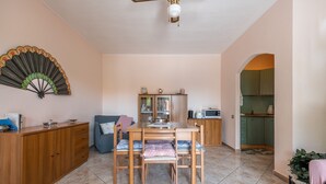 Un comedor con una mesa y sillas de madera, un ventilador en la pared y una cocina al fondo.