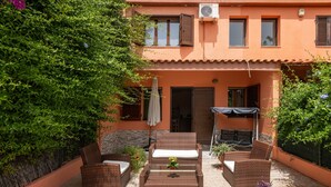 Un patio con mobiliario de mimbre, una mesa con un florero y un edificio de techo a tejas y ventanas.