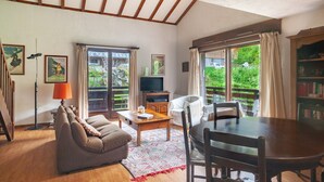A living room with a sofa, a coffee table, a TV, and a dining table with chairs. There are curtains, a bookshelf, and a window with a view of greenery. - Apartment at "Chalet Jade" with Mountain Views, Balcony, and Wi-Fi (Saint-Gervais-les-Bains)