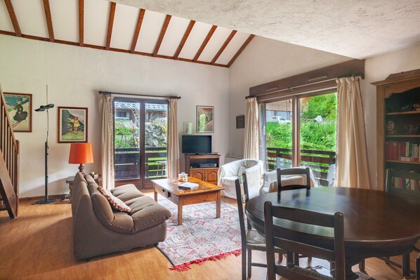 A living room with a sofa, a coffee table, a TV, and a dining table with chairs. There are curtains, a bookshelf, and a window with a view of greenery.