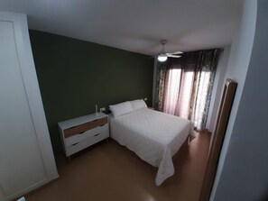 Een slaapkamer met een bed, nachtkastje, dressoir, plafondventilator en raam met gordijnen.