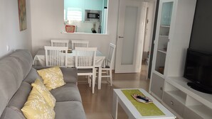 A living room with a grey sofa, white chairs, a coffee table, a TV, and a kitchen in the background. - Apartment 'Alegria' near the Beach with Shared Pool (Garrucha)