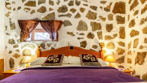 A bed with a purple blanket, two pillows with floral designs, a wooden headboard, stone walls, a window with brown curtains, and two bedside lamps. - Holiday Apartment "La Fortaleza - Casa La Fuentita - Adults Only" with Mountain Views (La Fuentita)
