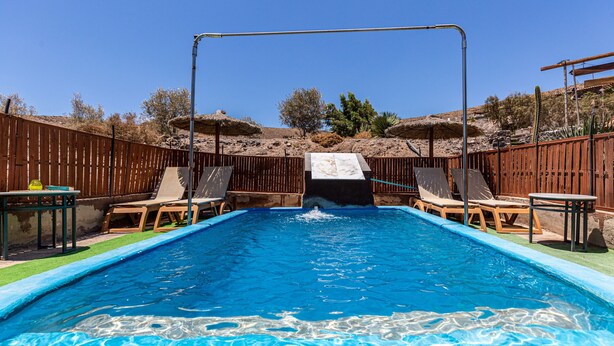 A rectangular swimming pool with a slide, surrounded by wooden fences, and equipped with lounge chairs and tables. - Holiday Apartment "La Fortaleza - Casa La Fuentita - Adults Only" with Mountain Views (La Fuentita)