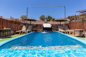 Piscina retangular com escorrega, cercada por vedação de madeira, e equipada com espreguiçadeiras e mesas.