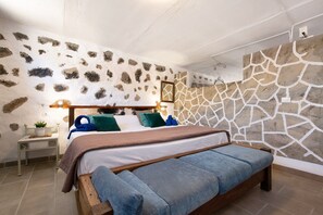 Un dormitorio con pared de piedra, una cama con almohadas azules y verdes, una mesita de noche con una planta y un banco de madera.