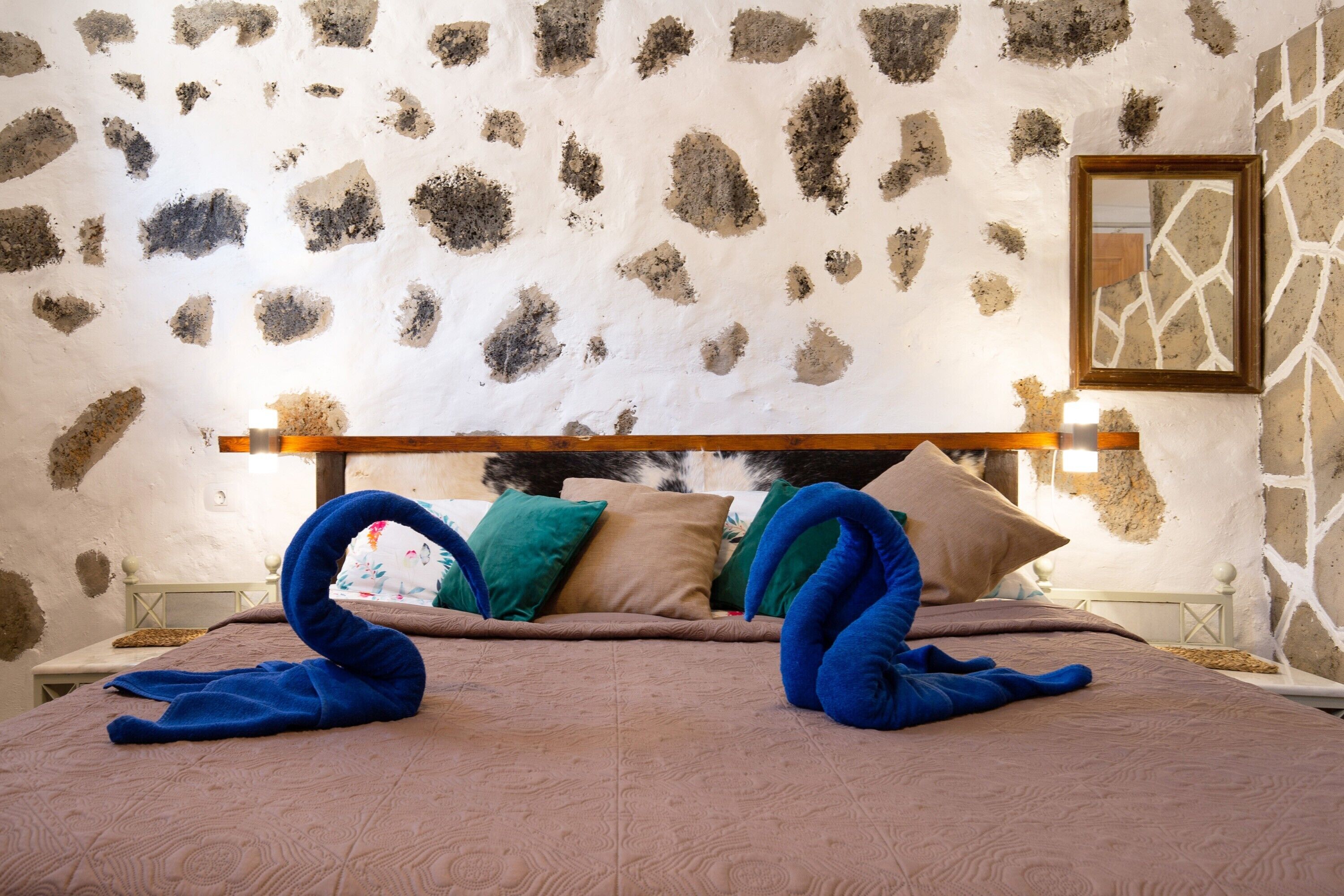 Un dormitorio con pared de piedra, estructura de cama de madera y cama con colcha marrón y almohadas azules.