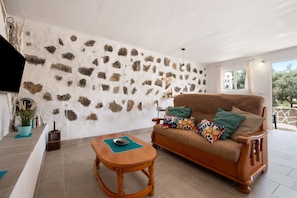 Ein Wohnzimmer mit einer Steinwand, einem Holzsofa mit bunten Kissen, einem Holztisch und einer Topfpflanze.