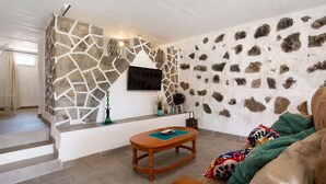 A living room with a stone wall, a mounted TV, a wooden table, and a sofa with colorful blankets. - Holiday Home 'Esencia Eco-rústico - Adults Only' with Shared Pool and Garden (La Fuentita)