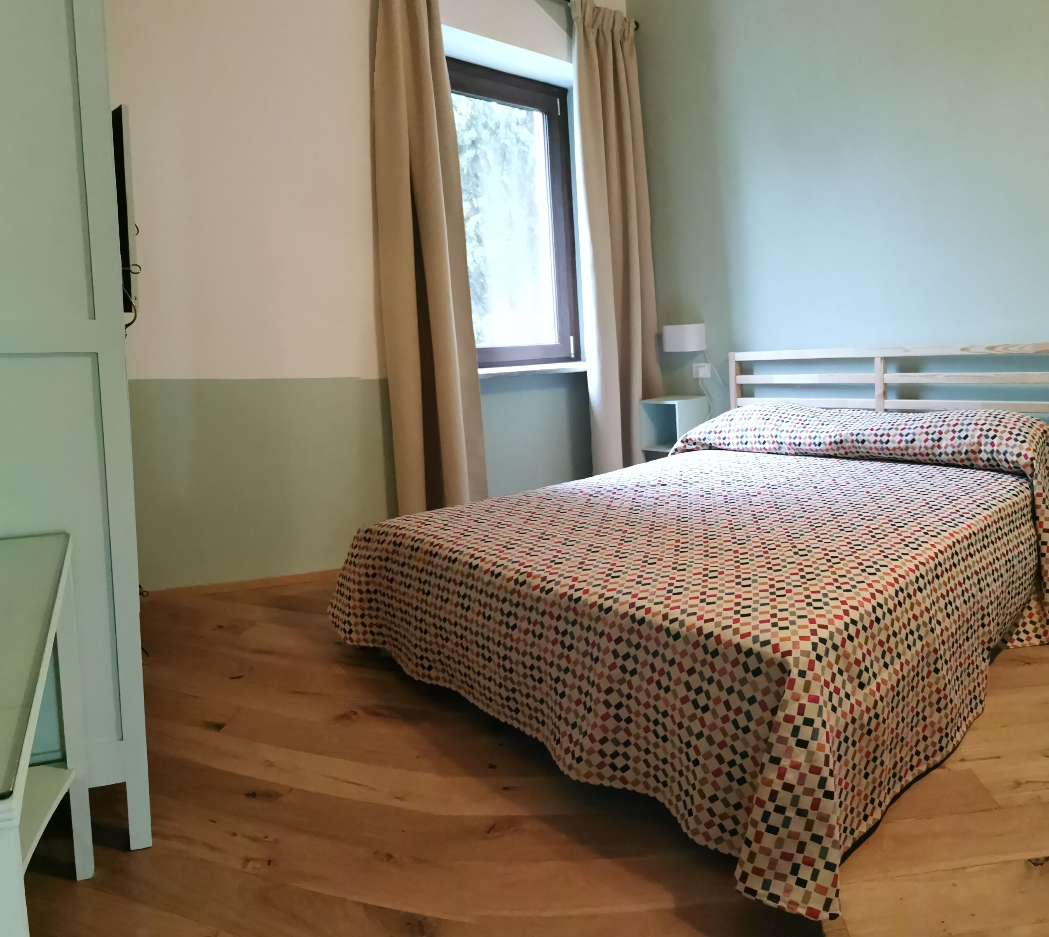 Ein Schlafzimmer mit Holzboden, einem Bett mit gemustertem Überwurf, einem Fenster mit Vorhängen und einem Nachttisch mit Lampe.