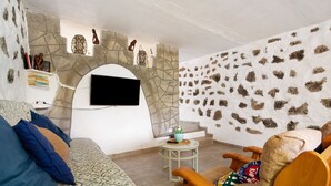 A living room with a stone wall, a mounted TV, a sofa, a chair, and a small table with a plant. - Adults-Only Holiday Home Alma Eco-Rustic with Shared Pool and Terrace (La Fuentita)