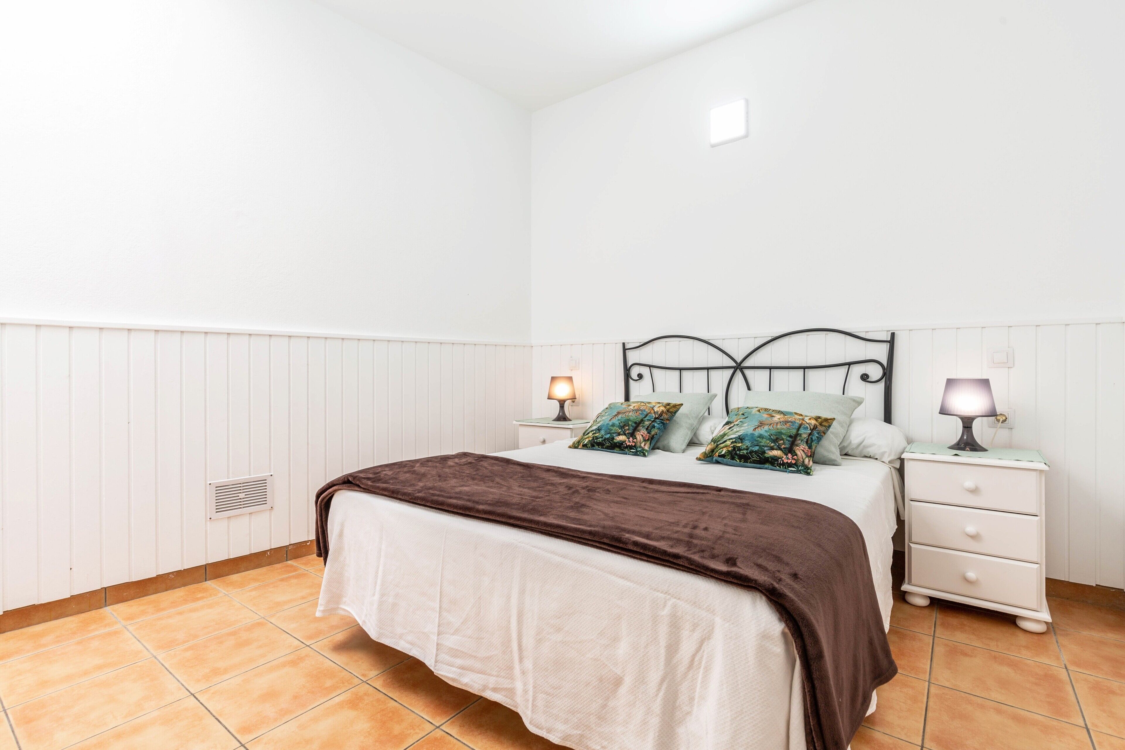 Un dormitorio con una cama, dos mesitas de noche con lámparas, un calentador y una pared blanca.