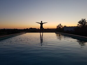 A person standing in a swimming pool with arms outstretched, surrounded by calm water and a sunset sky.