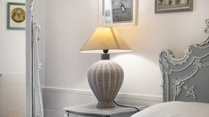 Una mesita de noche blanca con una lámpara de mimbre, una cama blanca con cabecero decorativo, y un cuadro en la pared.