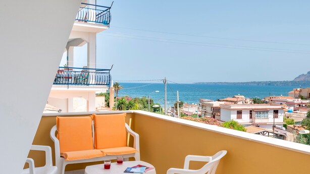 Ein Balkon mit weißen Plastikstühlen und einem Tisch, einer gelben Wand und Blick auf das Meer und Gebäude.